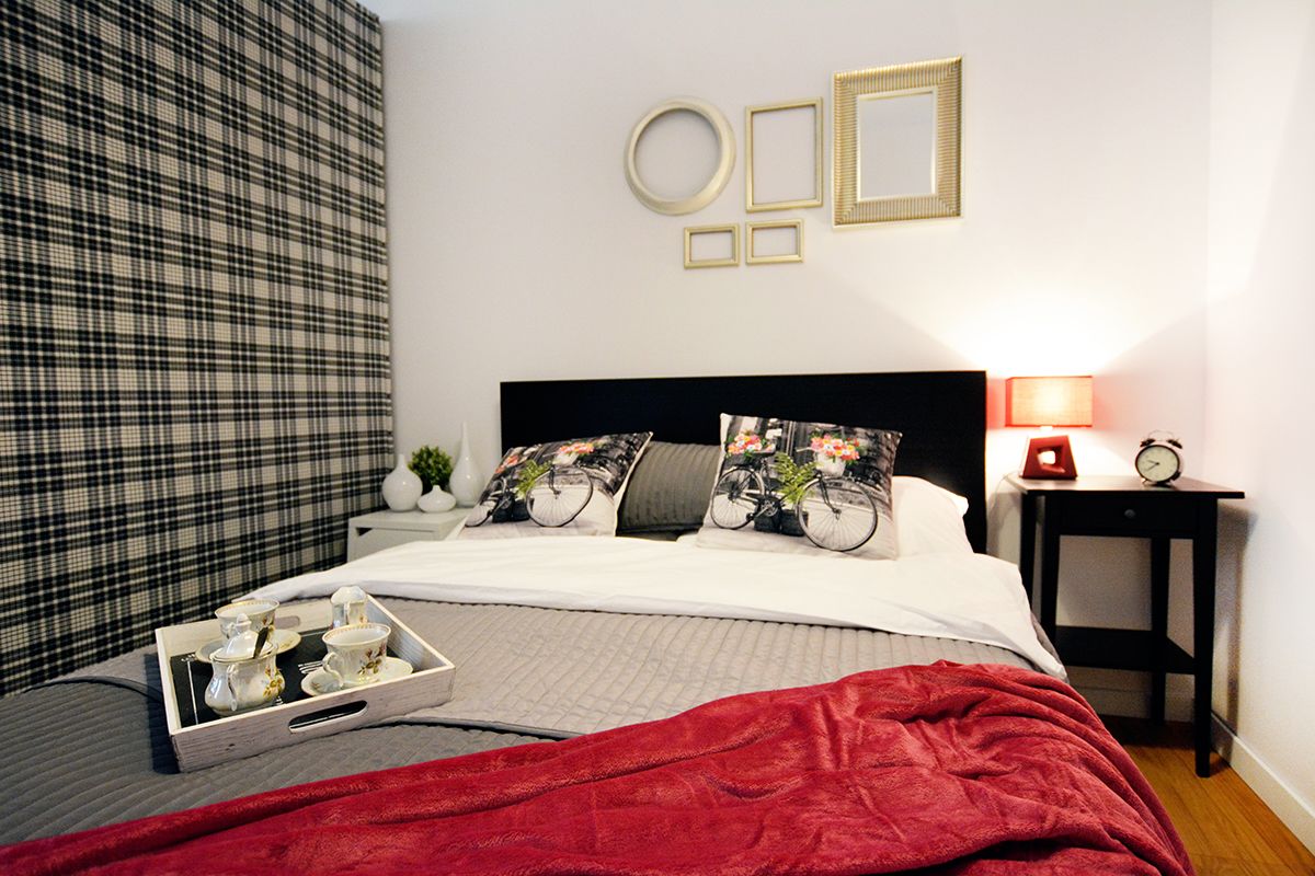 Een net opgemaakt bed met een rode deken, een nachtkastje met een lamp en een muur met ingekaderde foto's.
