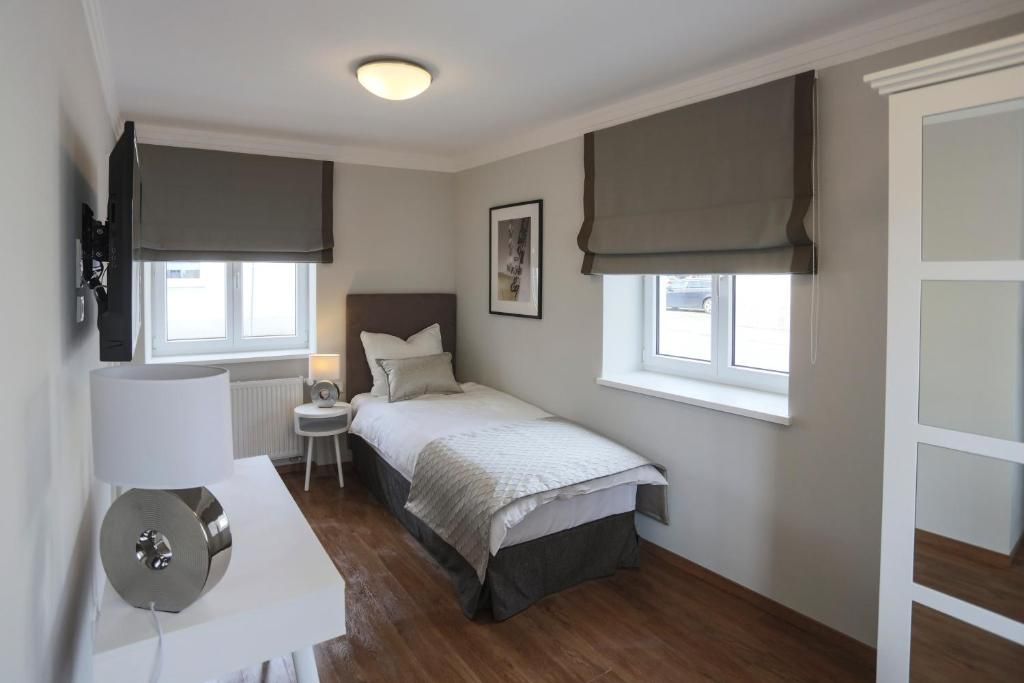 Quarto com cama, mesa de cabeceira, abajur e janela com cortinas.