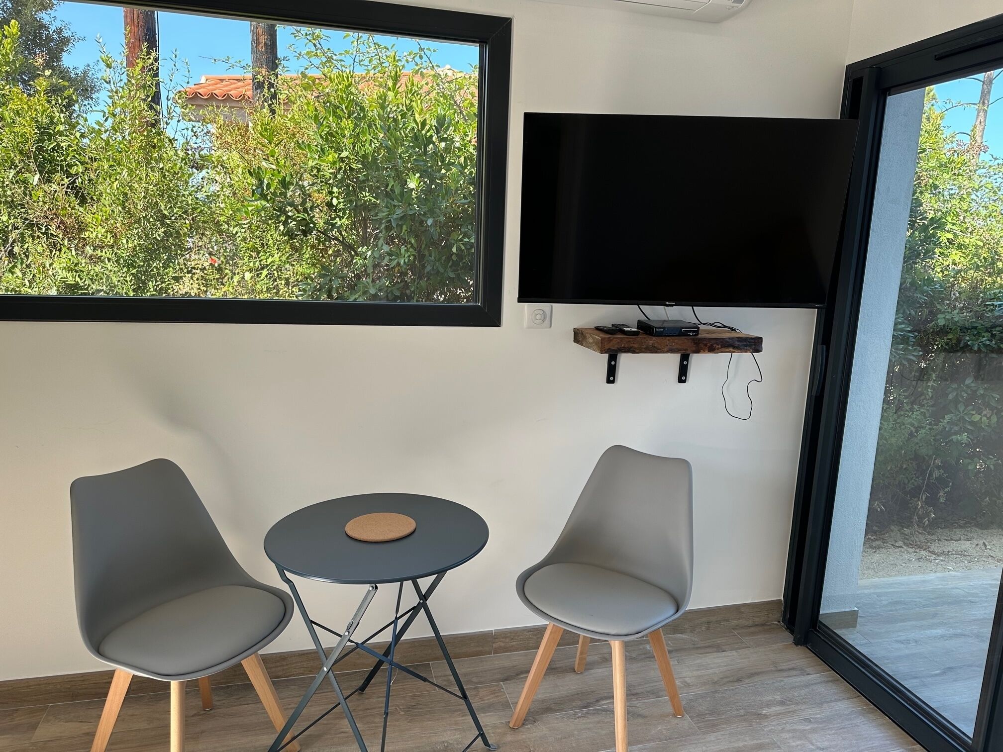 Ein modernes Zimmer mit großem Fenster, einem an der Wand montierten Flachbildfernseher und zwei grauen Sesseln um einem kleinen runden Tisch.