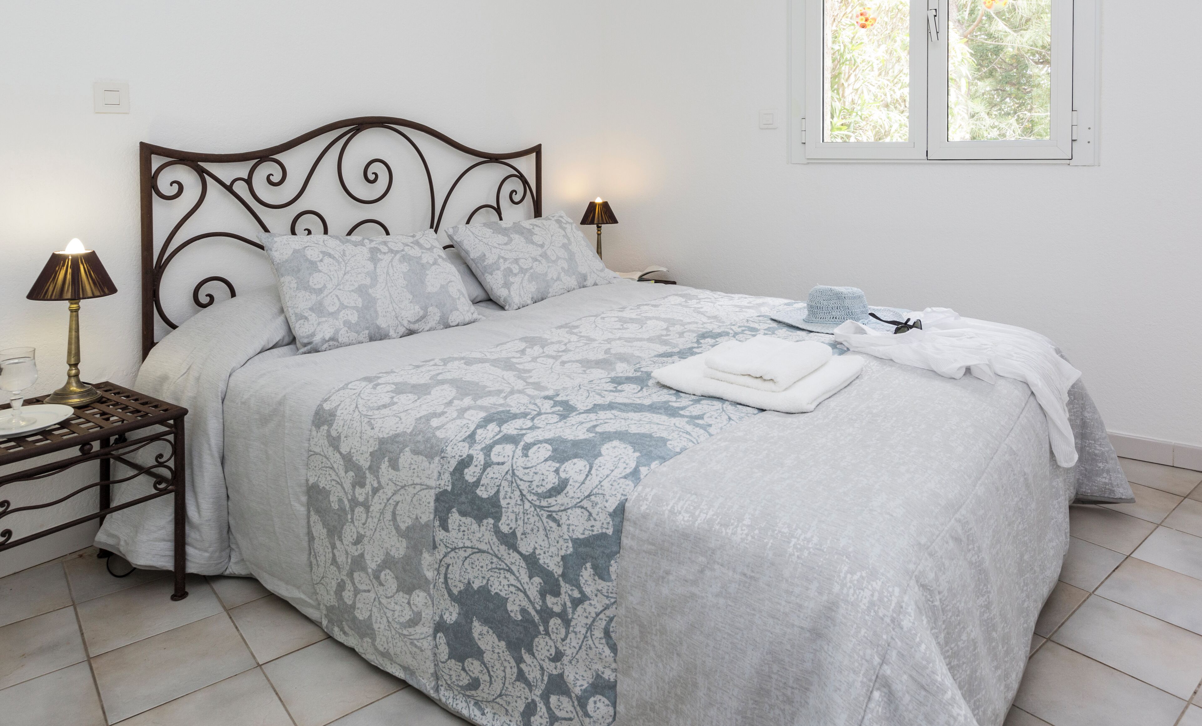 Una cama bien tendida con una colcha estampada, una mesita de noche con una lámpara, y una ventana que da a zonas verdes.