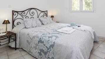 Una cama bien tendida con una colcha estampada, una mesita de noche con una lámpara, y una ventana que da a zonas verdes.
