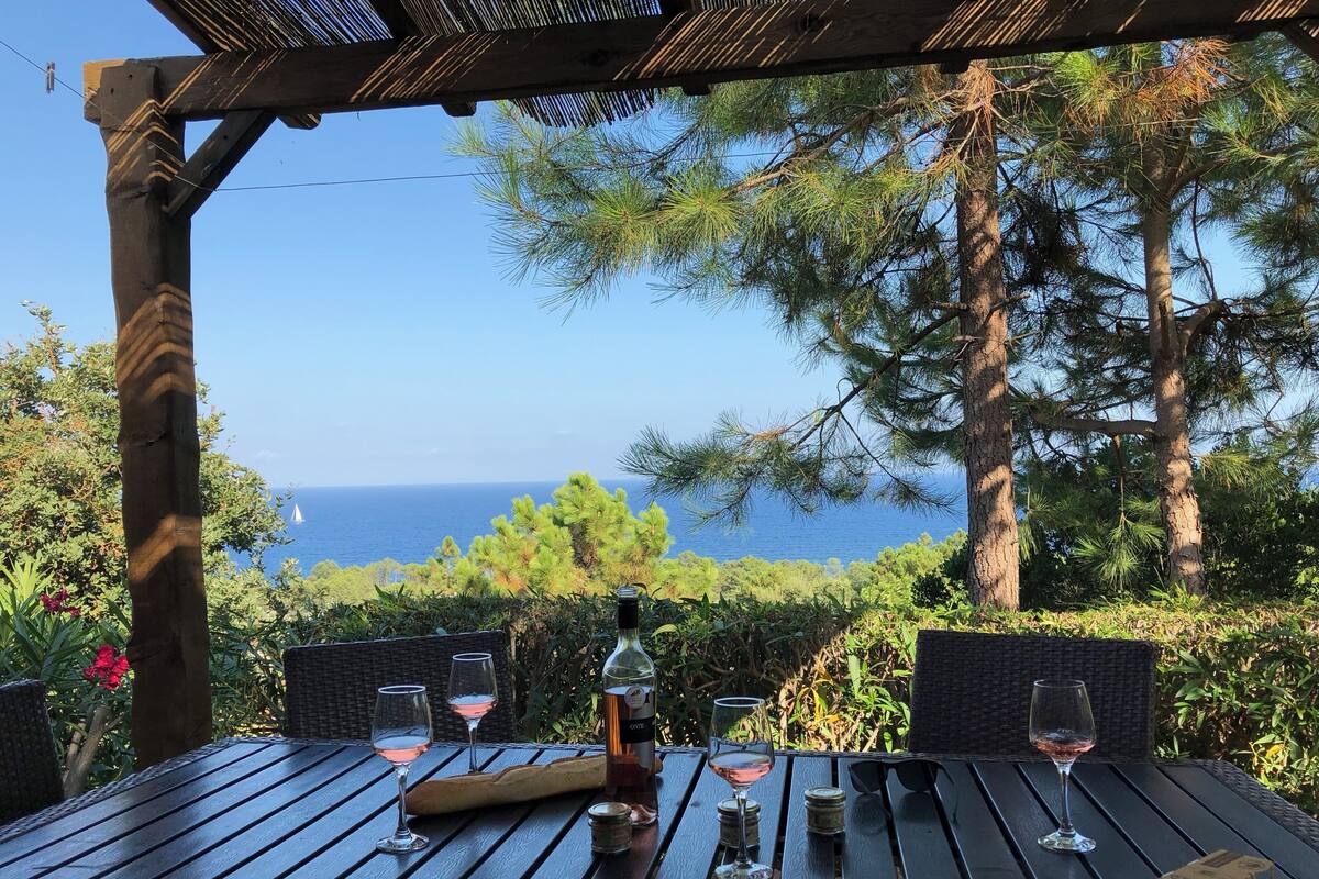 Ein Essbereich im Freien mit Meerblick, ausgestattet mit einer Holzhütte, Korbstühlen und einem Tisch gedeckt mit Weingläsern und einer Weinflasche.