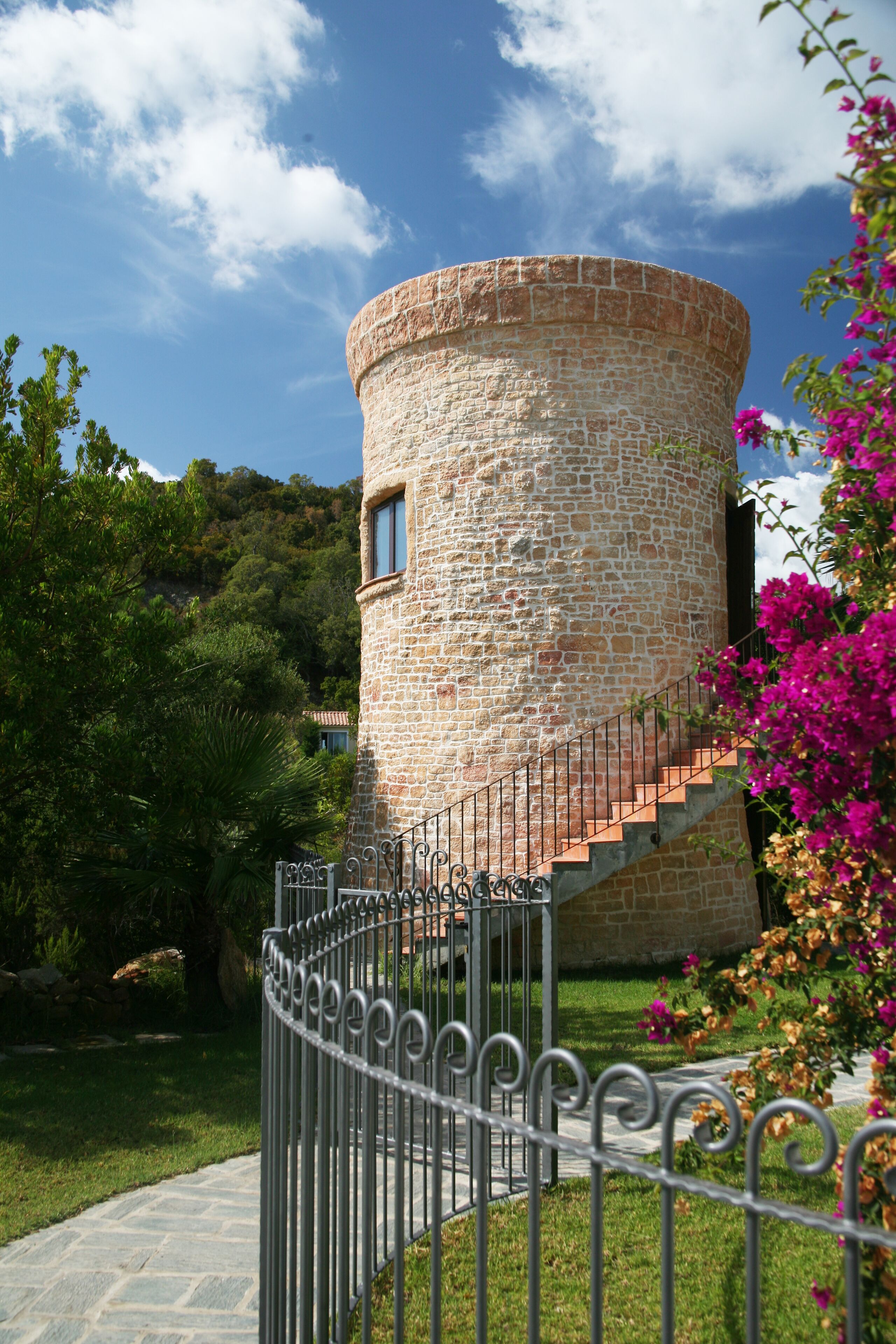 A stone tower with a spiral staircase leading to a window, surrounded by a metal fence and lush greenery.