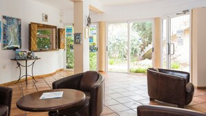 A spacious living area with a large sliding glass door leading to a garden, a brown leather sofa, a small round table, and a wooden coffee table. - Mare e Monte (Sari-Solenzara)