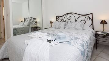 Een net opgemaakt bed met een beddekleed met patroon, een witte handdoek en een witte hoed. Er staan twee nachtkastjes met lampen, een spiegel en een deur die naar een andere kamer leidt.