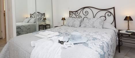 A neatly made bed with a patterned bedspread, a white towel, and a white hat on the bed. There are two bedside tables with lamps, a mirror, and a door leading to another room.