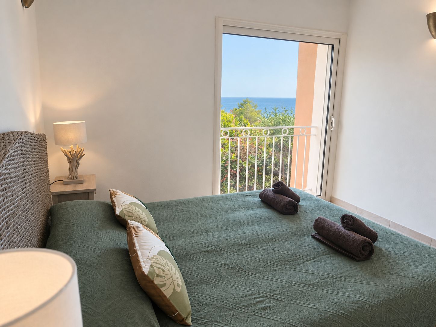 Un dormitorio con colcha verde, dos almohadas, una mesita de noche con lámpara y vistas al mar a través de una puerta de balcón.