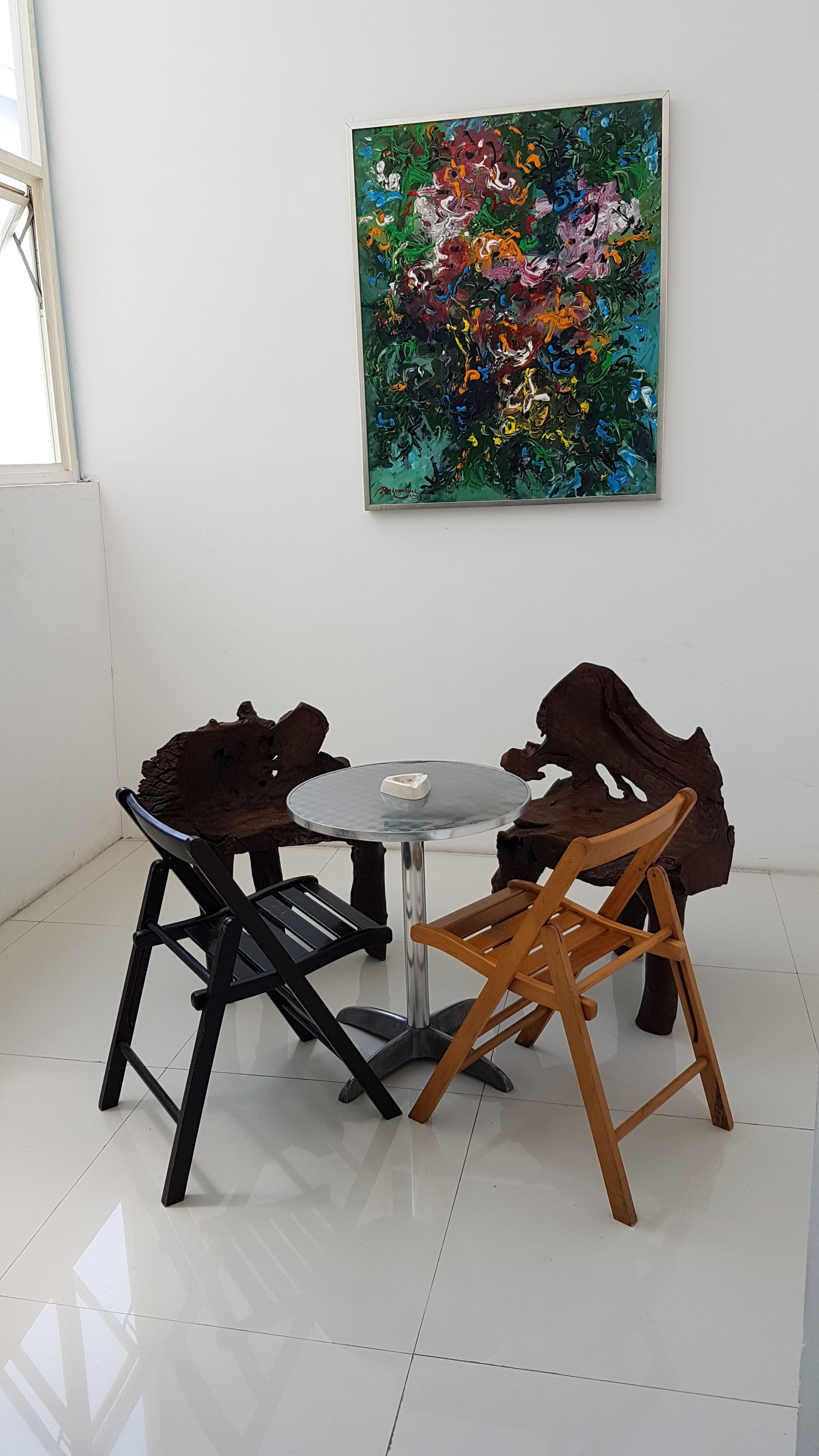 Un pequeño comedor con una mesa redonda y dos sillas, una negra y otra de madera, en una habitación con paredes blancas y un cuadro abstracto de colores sobre la mesa.