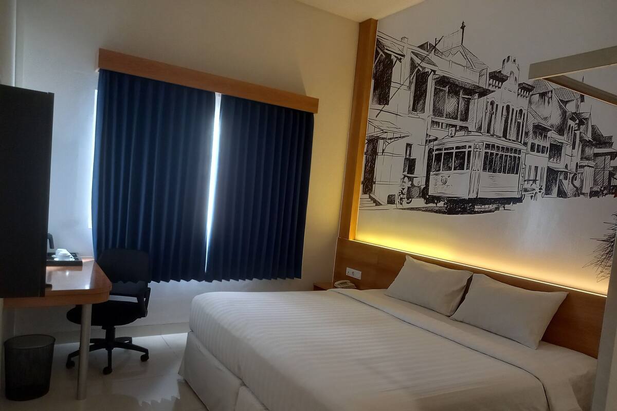 A hotel room with a bed, a desk with a chair, a trash bin, a window with curtains, and a wall mural.