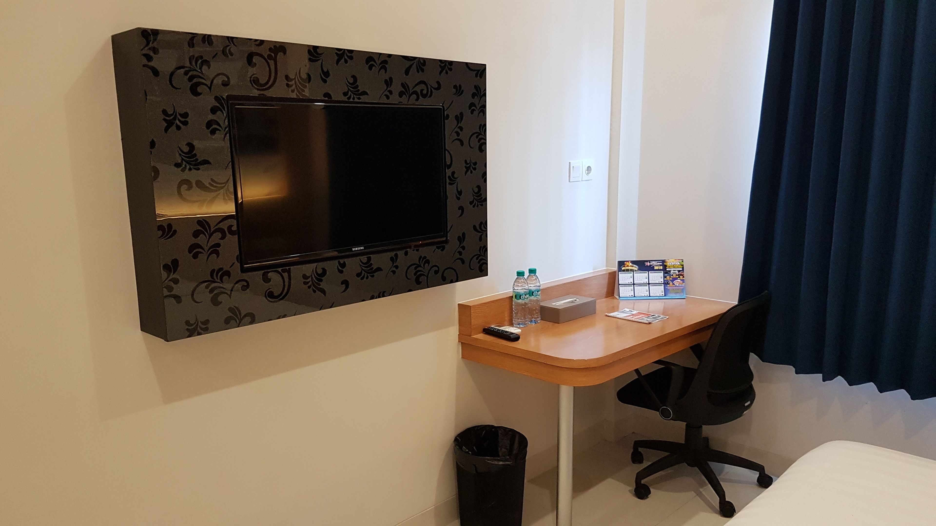 Une chambre d’hôtel équipée d’un téléviseur fixé au mur, d’un bureau en bois avec une chaise, d’une poubelle et d’un calendrier accroché au mur.