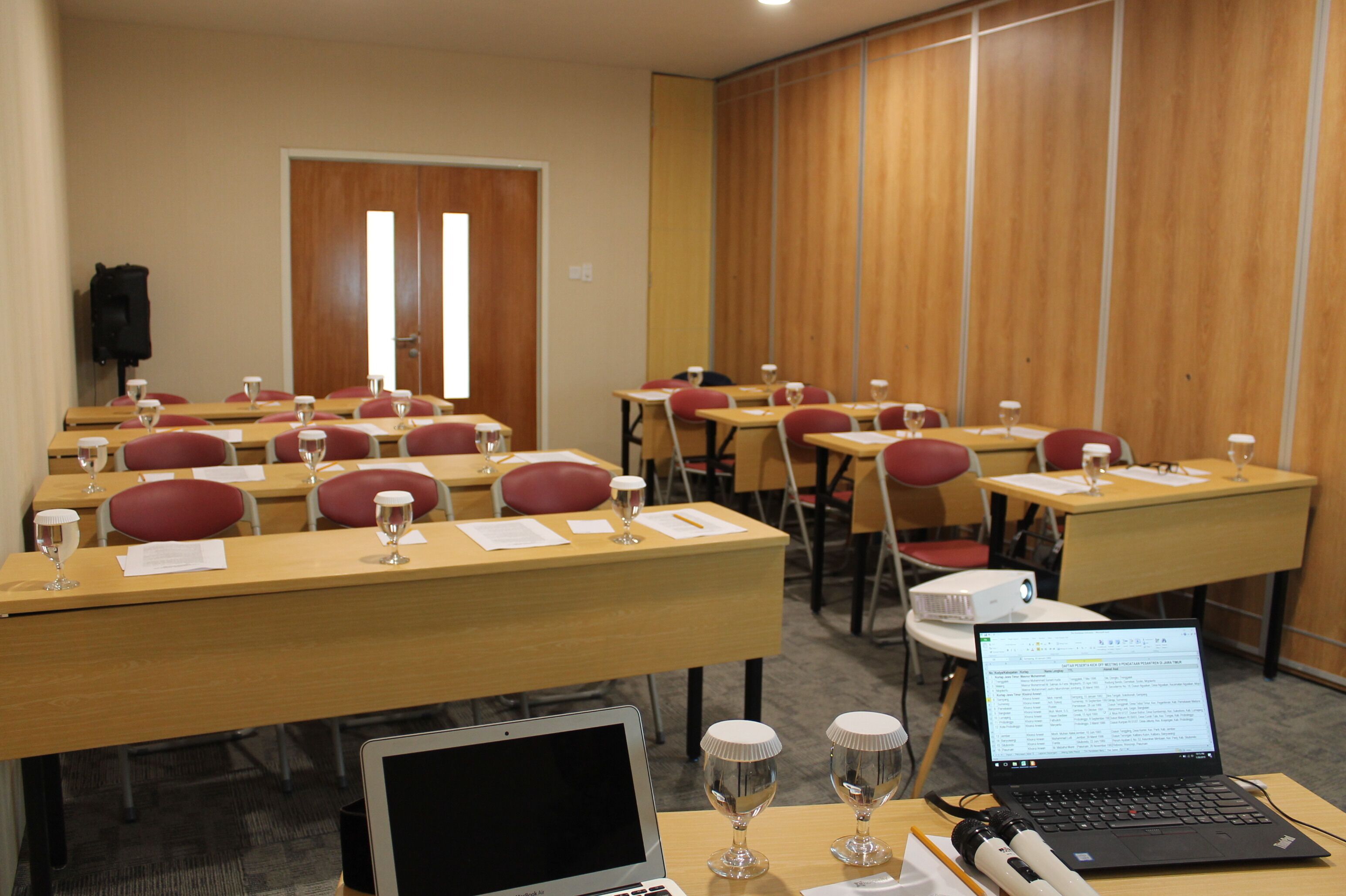 Un salón de conferencias con filas de escritorios, cada uno con una computadora portátil, documentos y un vaso de agua.