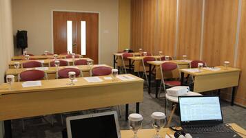 Un salón de conferencias con filas de escritorios, cada uno con una computadora portátil, documentos y un vaso de agua.