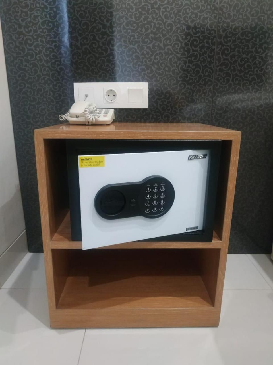 A safe with a keypad, placed in a wooden cabinet with a power outlet above it.