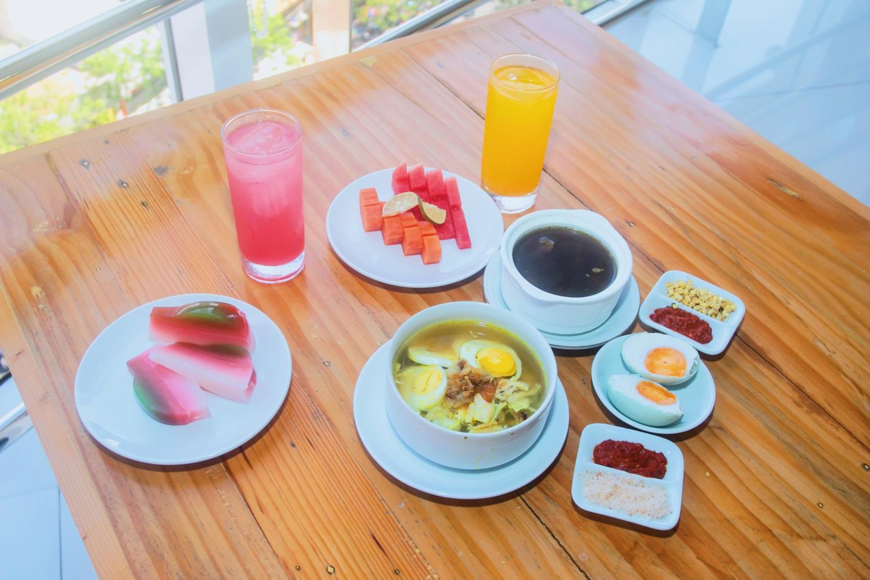 Una mesa de madera con una comida que incluye una bebida rosa, sandía, un tazón de sopa con huevos y diferentes salsas.