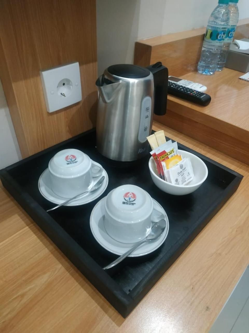 A hotel room's breakfast setup with a kettle, two cups, a bowl of cereal, and a remote control on the counter.