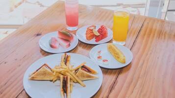 Una mesa de madera con una fuente de sándwiches de queso a la plancha, papas fritas, rebanadas de sandía y un plátano. También hay dos bebidas, una rosa y otra naranja.