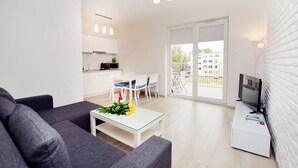 Uma sala moderna com sofá, um espaço de jantar com cadeiras e mesa, uma cozinha com armários e bancada, e uma varanda com televisão.