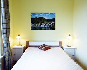 A hotel room with a bed, two bedside tables with lamps, a painting of a waterfall, and a curtain. - Golden Sun Apartments- Osiedle Pogodne (Kolobrzeg)
