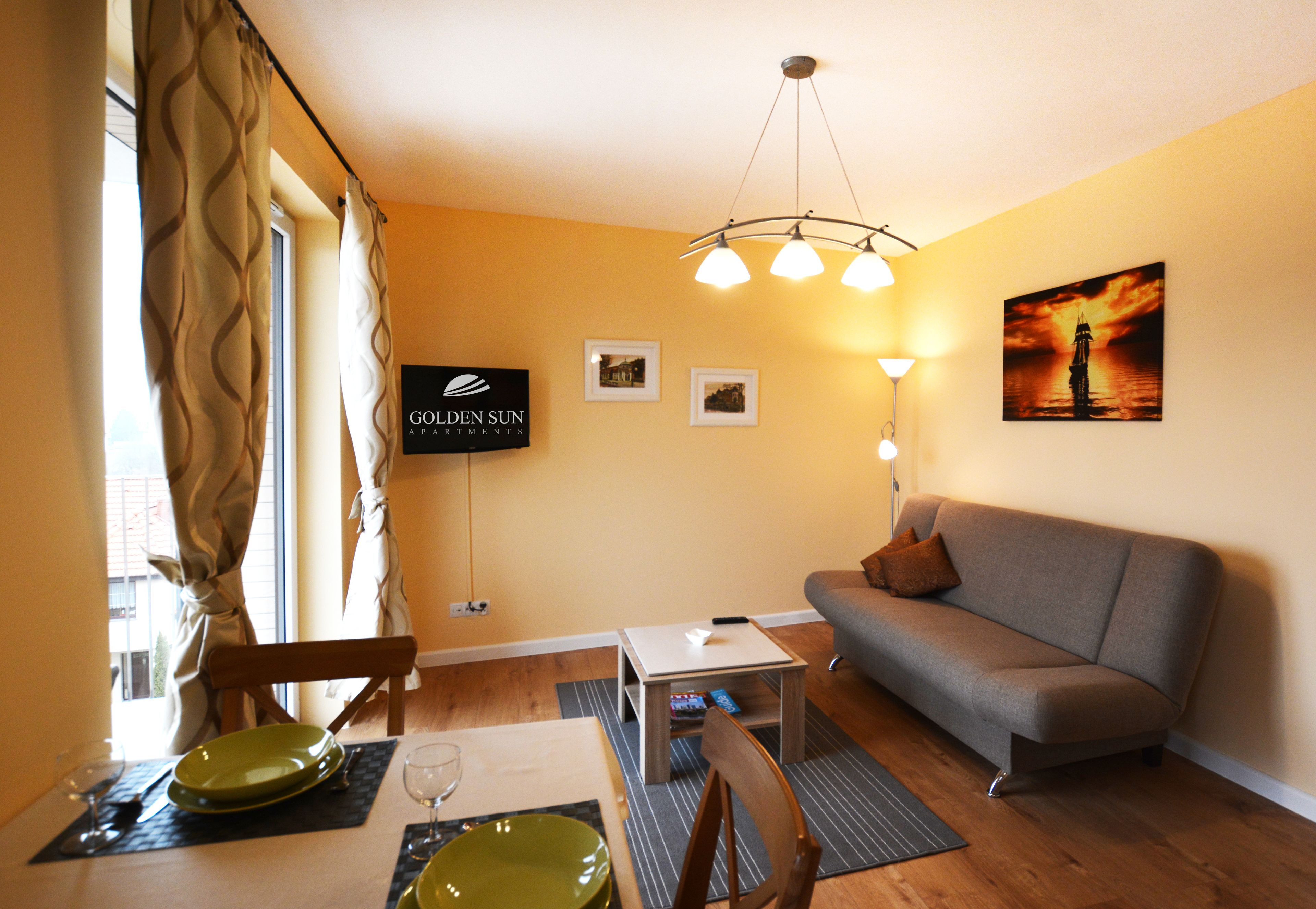 A living room with a sofa, a dining table with plates and glasses, a small side table, and a wall-mounted TV displaying the logo "GOLDEN SUN APARTMENTS".