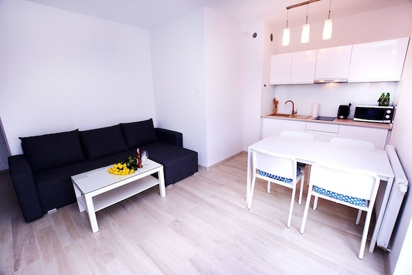 Un espace de vie compact comprenant un canapé noir, une petite table blanche agrémentée d’un arrangement floral, une kitchenette équipée d’un four à micro-ondes et un coin repas avec une table et des chaises blanches.