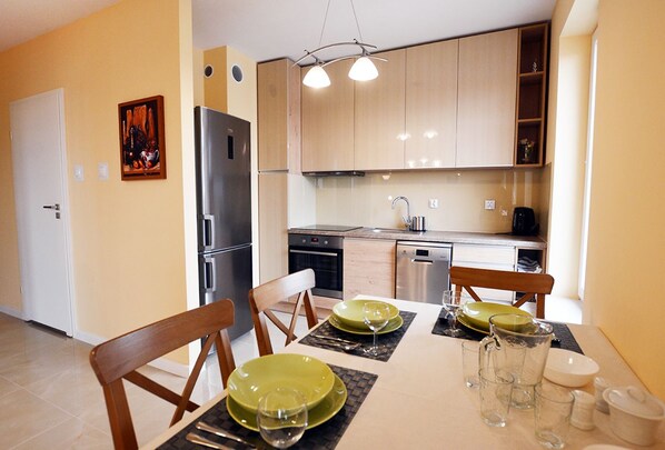 A modern kitchen with wooden cabinets, a stainless steel refrigerator, and a dining area with a table set for four. - Golden Sun Apartments- Osiedle Pogodne (Kolobrzeg)