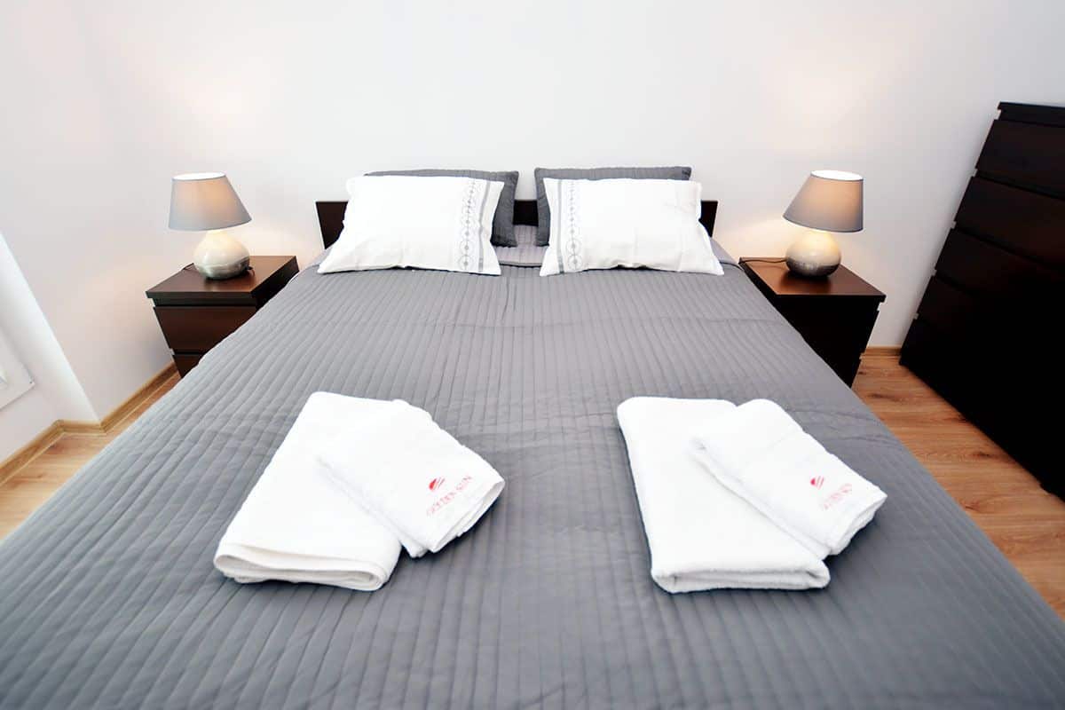A neatly made bed with a grey quilt, two white pillows, and two folded towels on the bed. There are two bedside tables with lamps, and a dark headboard.