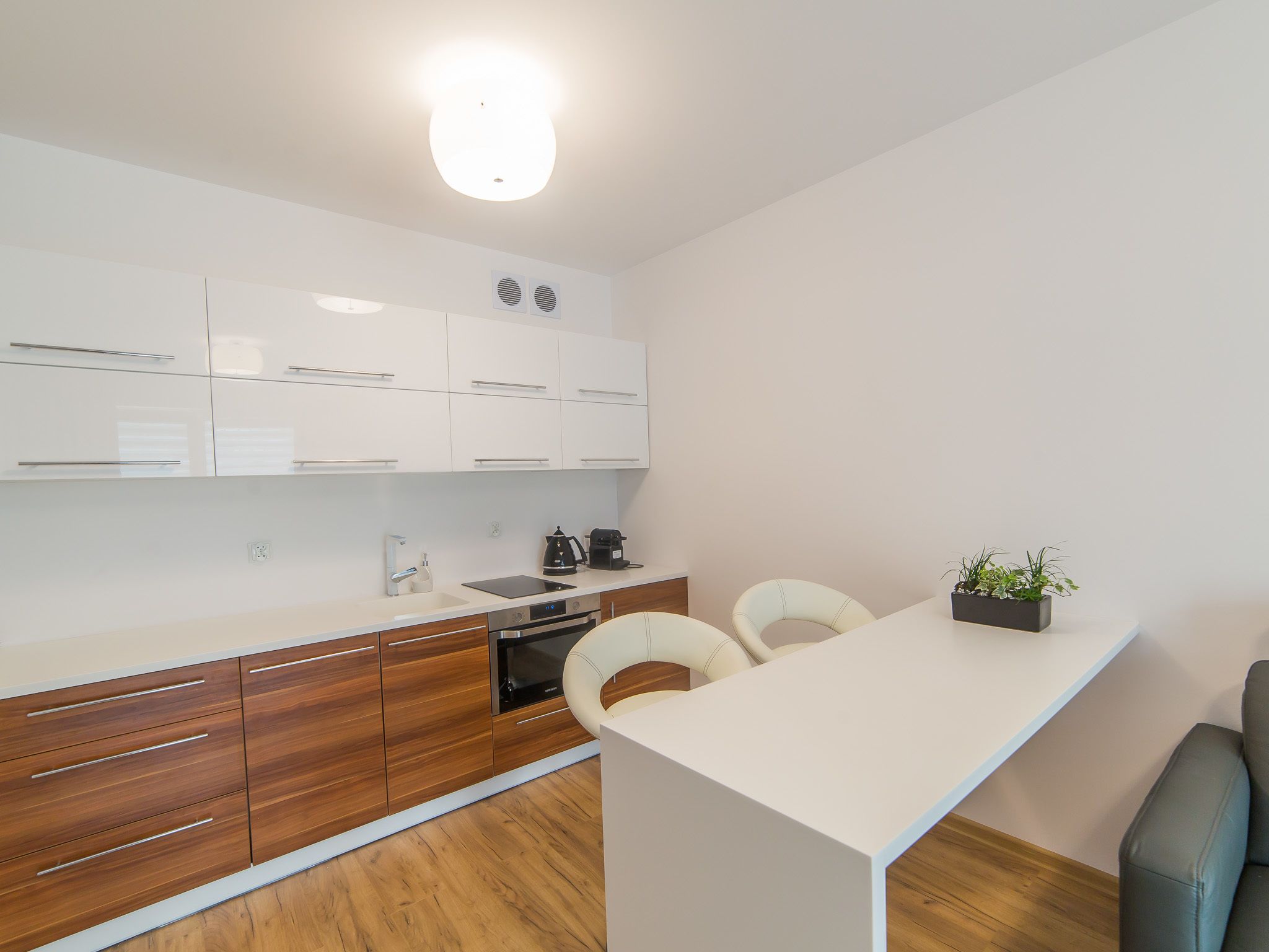 Eine moderne Küche mit weißen Schränken, einer Holz Arbeitsplatte und einem eingebauten Backofen. Auf der Arbeitsplatte stehen zwei Stühle und ein kleiner Topf mit Pflanze.
