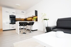 A modern kitchen with white cabinets, a black countertop, and a bar area with stools. There is a white vase with flowers on a table, and a black sofa in the living area.