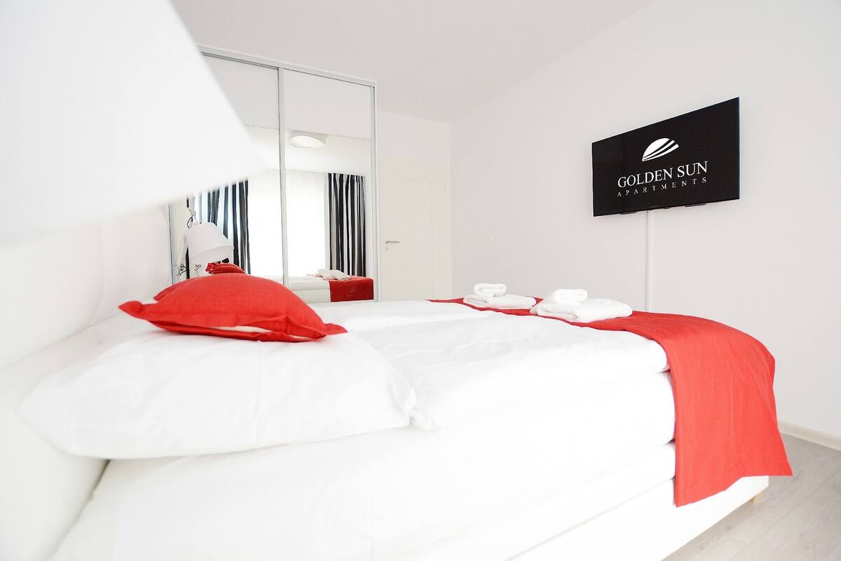 A modern hotel room with a large bed, a red throw pillow, white bedding, a mirror, and a sign for Golden Sun Apartments.