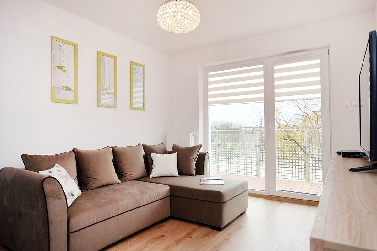 A modern living room with a brown sectional sofa, a flat-screen TV, and a large window with blinds.