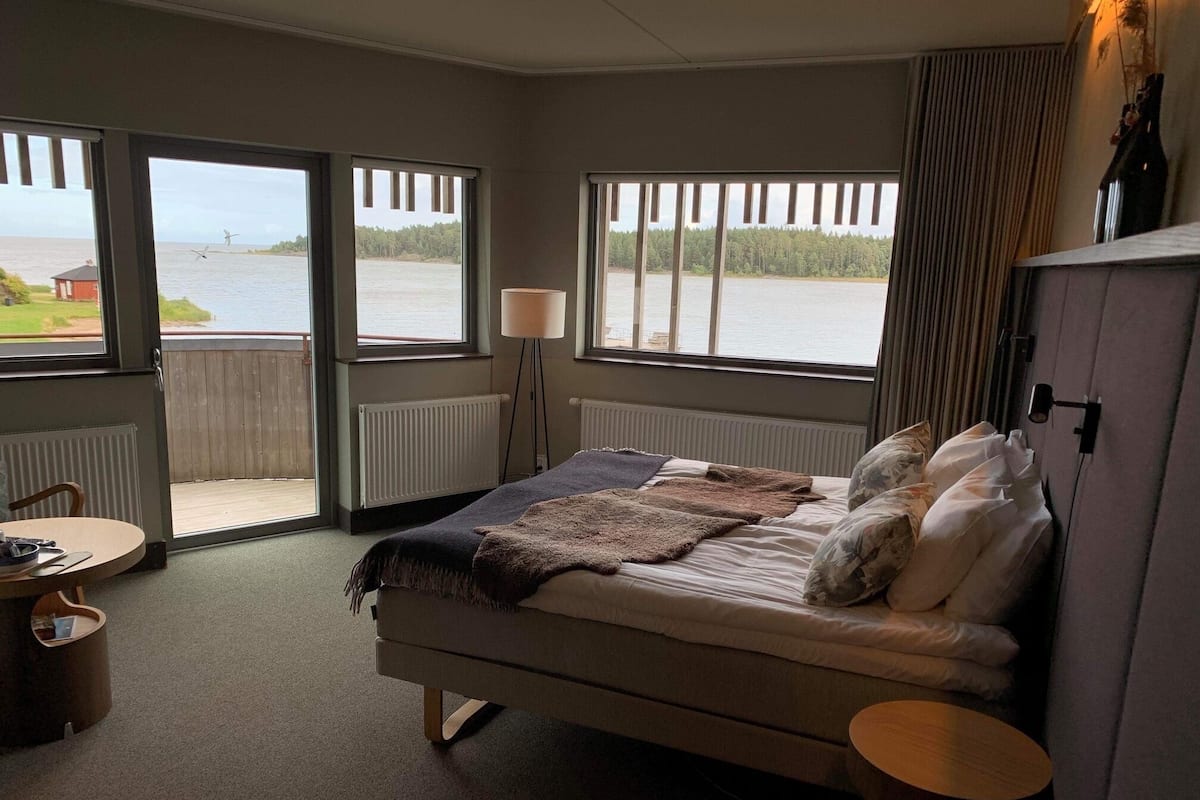 Un dormitorio con cama, mesita de noche, lámpara, vista hacia el agua y ventana con persianas.