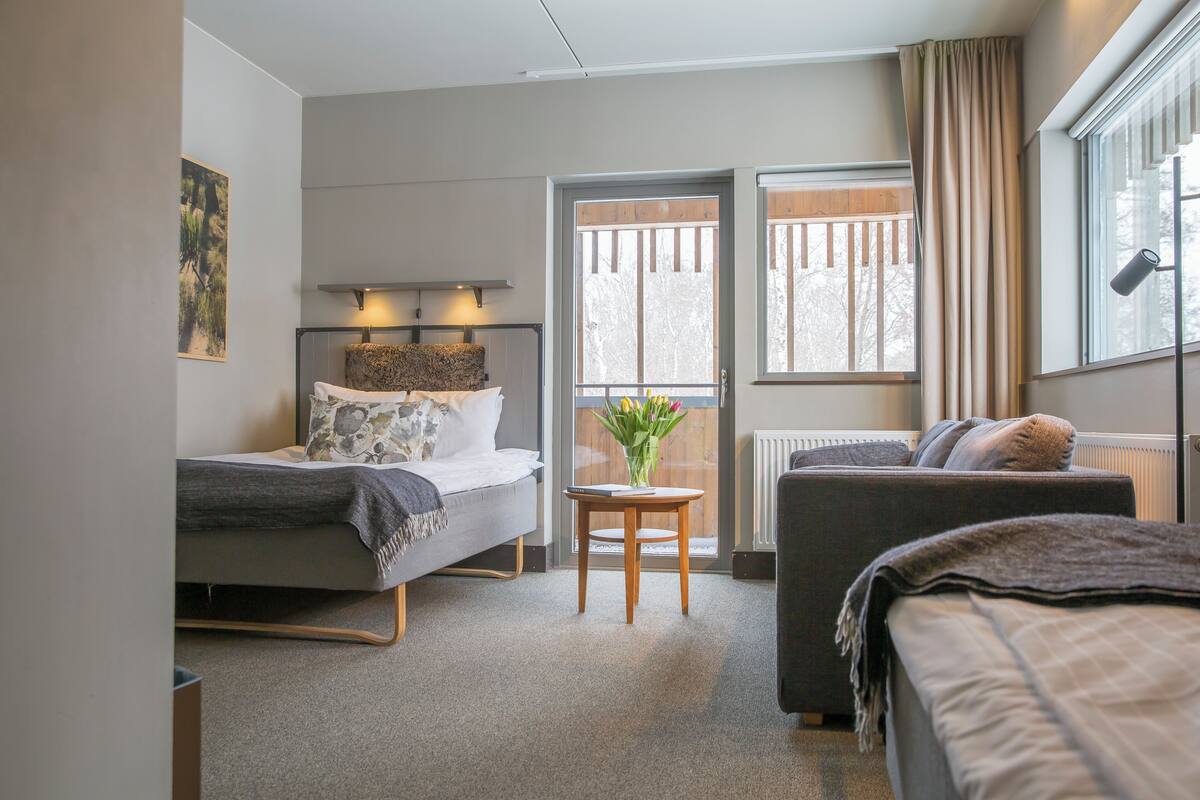 Una habitación de hotel moderna con cama, sofá, una mesita con un florero y ventana con cortinas.