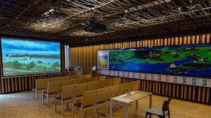 Uma sala de conferências com cadeiras de madeira, uma tela de projetor exibindo um mapa e uma parede revestida em madeira.