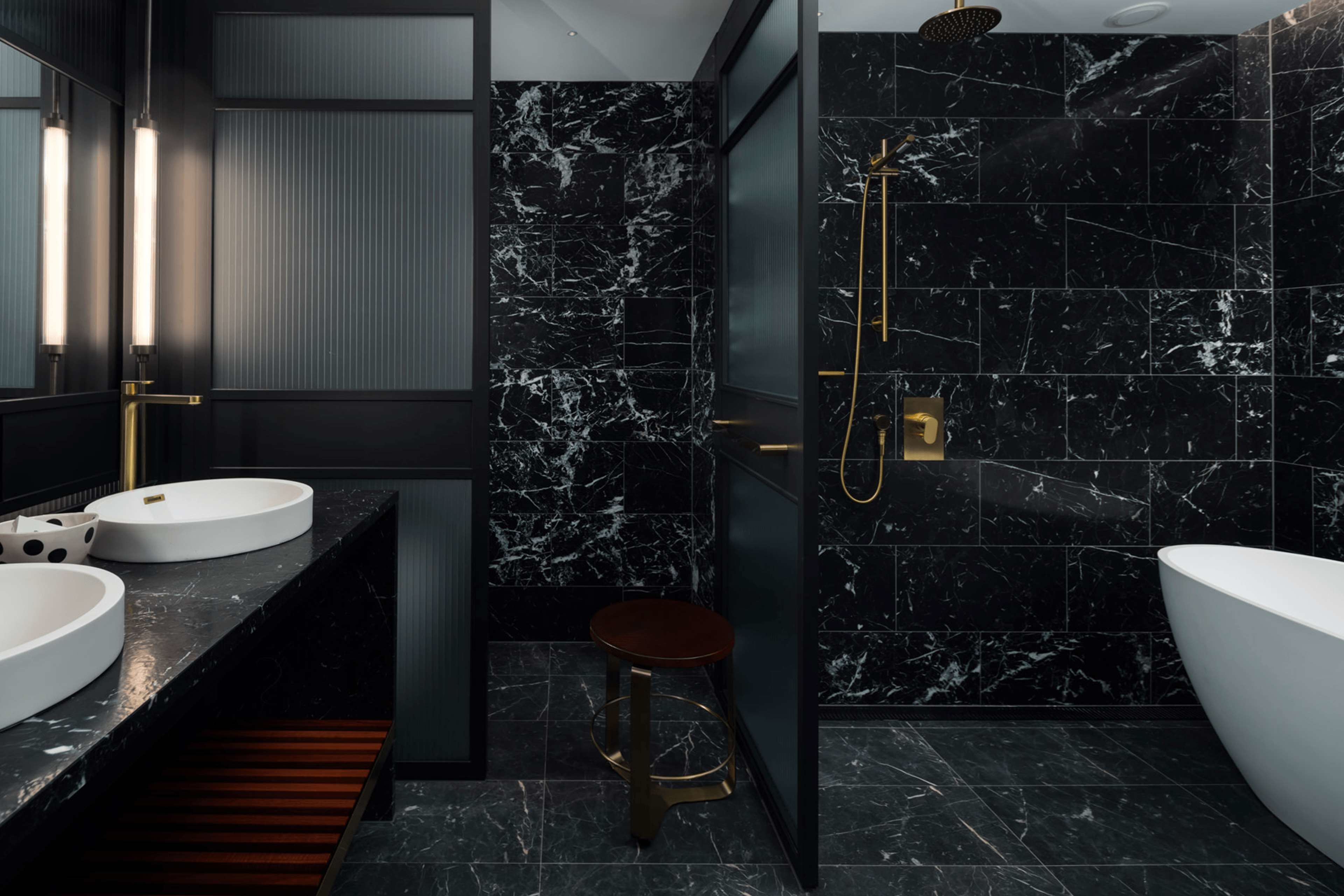 Un baño moderno con baldosas de mármol negro, un lavamanos blanco, una bañera grande y un cabezal de ducha dorado.