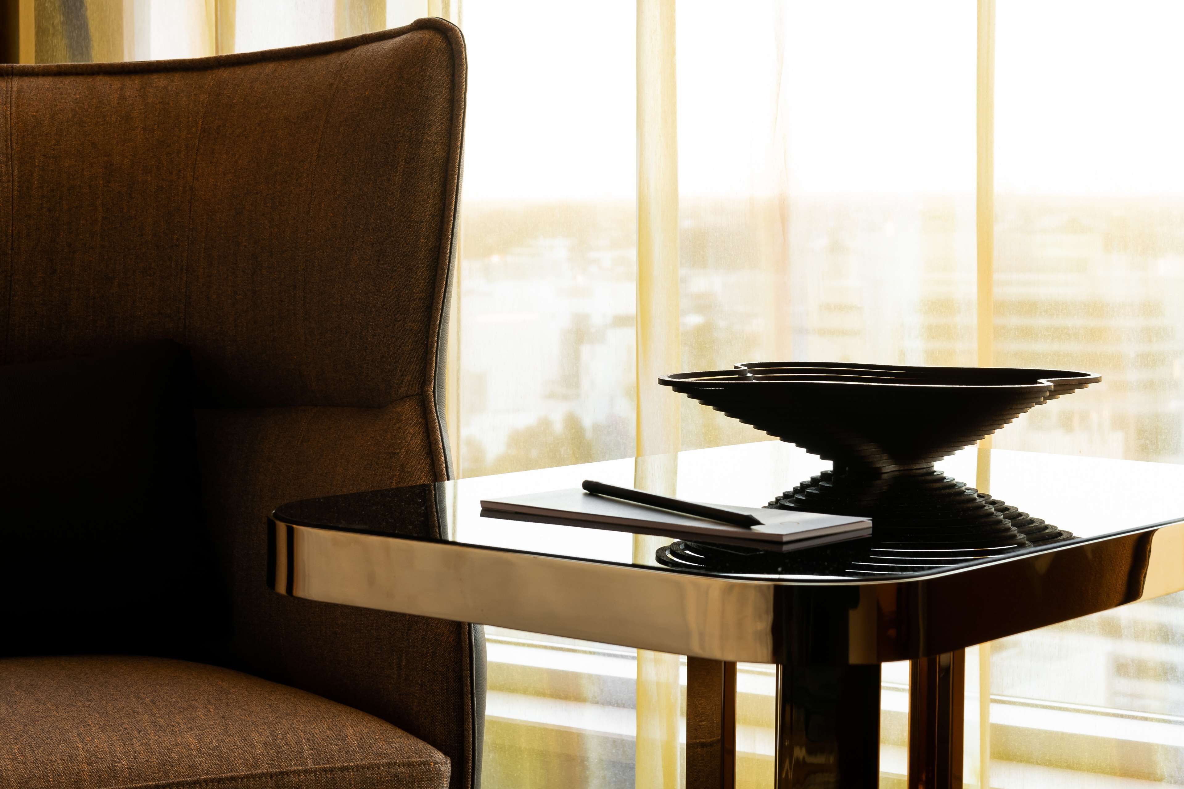 A modern room with a dark brown armchair, a small table with a black object, and a view of a cityscape through large windows.
