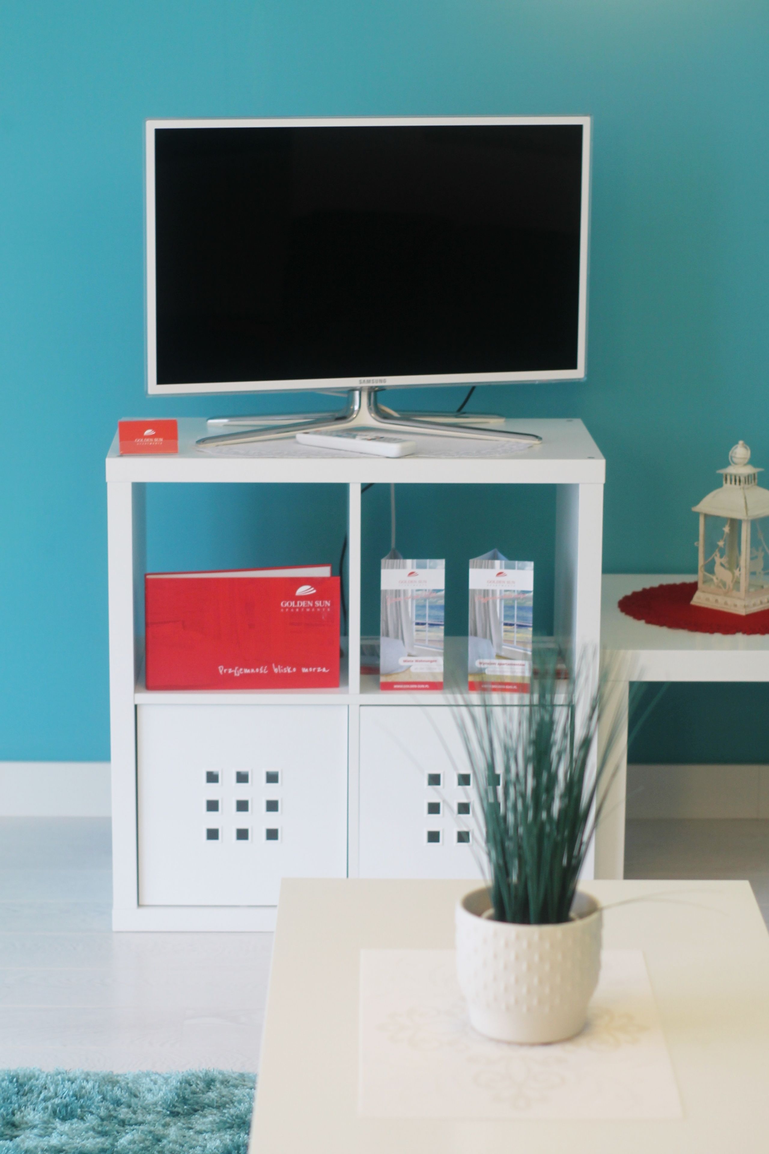 Un mueble de entretenimiento blanco con un televisor de pantalla plana, una caja roja con la etiqueta “GOLDEN SUN” y una pequeña planta en maceta.