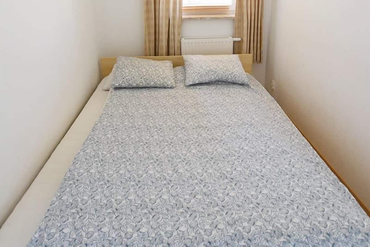 A single bed with a patterned bedspread, beige curtains, a window with blinds, and a ceiling light fixture.