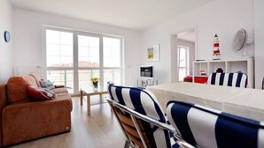 A modern living room with a sofa, dining table, and chairs. There is a large window providing natural light and a view of the outdoors. - Golden Sun Apartments  - Grzybowo (Kolobrzeg)