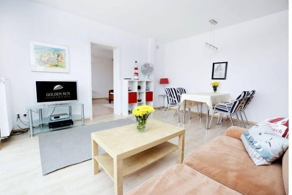 A modern living room with a flat-screen TV, a coffee table with yellow flowers, a sofa, and a dining area with chairs and a table.