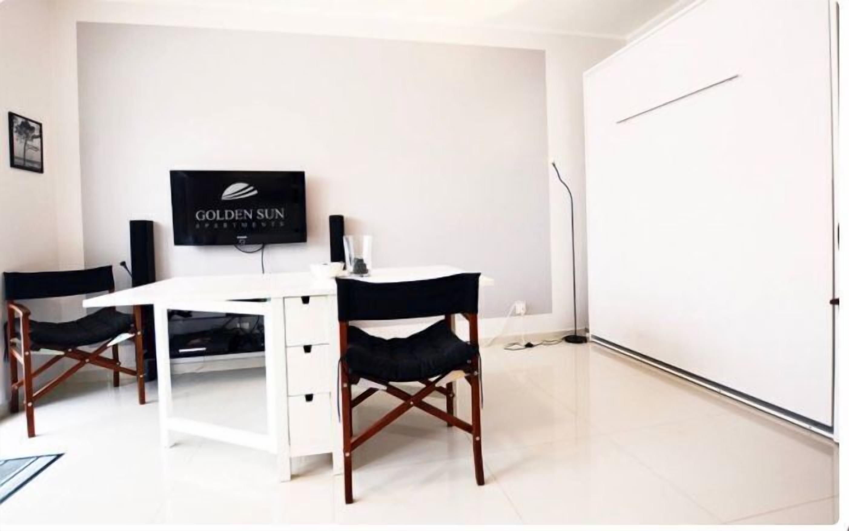 A room with a white desk, two black chairs, a flat-screen TV on the wall, and a closed door.