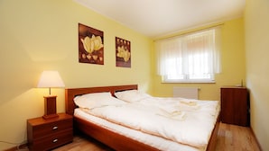 Un dormitorio con cama de madera, una mesita de noche con lámpara, dos cuadros colgados en la pared, una ventana con cortinas y un radiador.