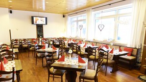 Un restaurante con varias mesas y sillas, un televisor montado en la pared y amplios ventanales con cortinas.