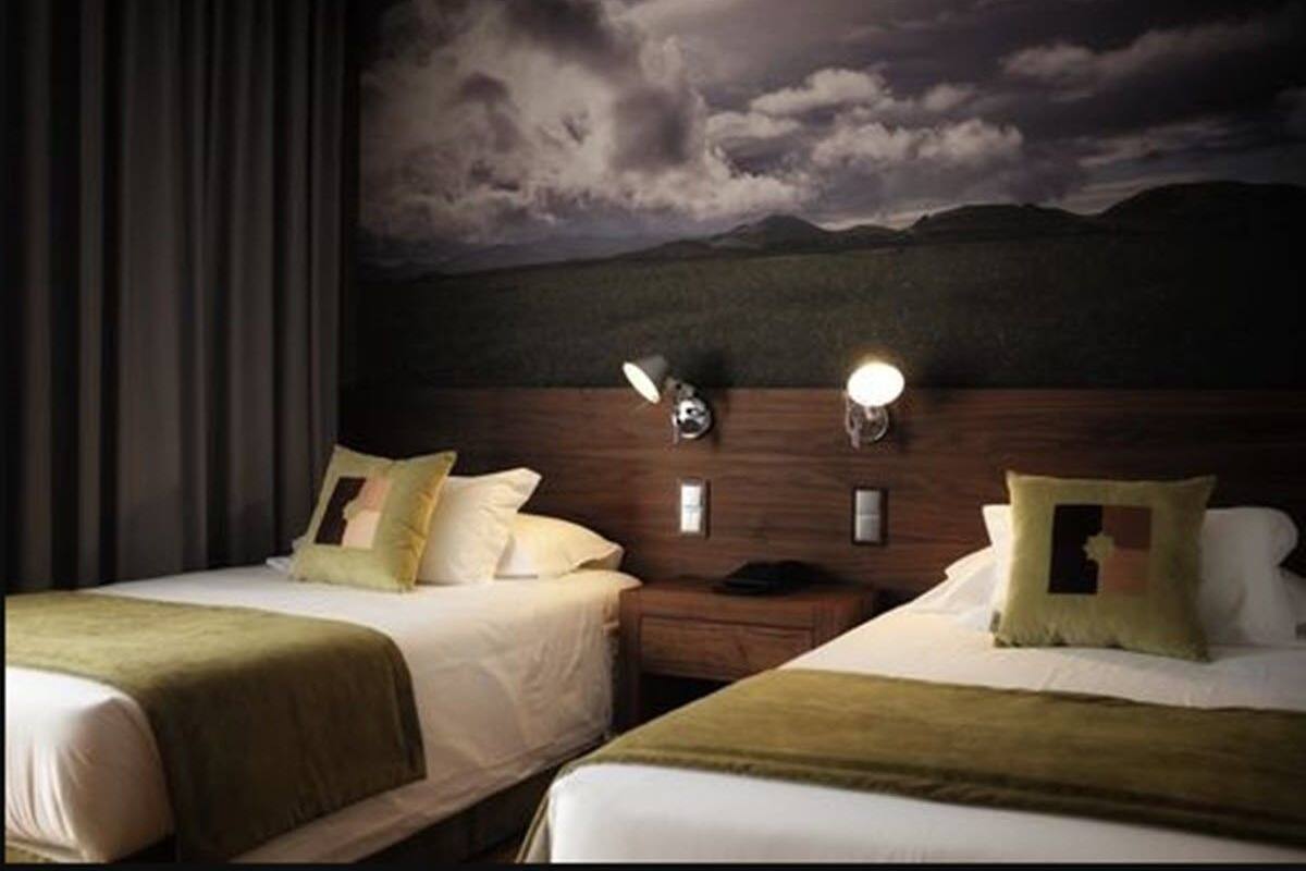 A hotel room with two beds, a nightstand, a lamp, and a wall-mounted light.