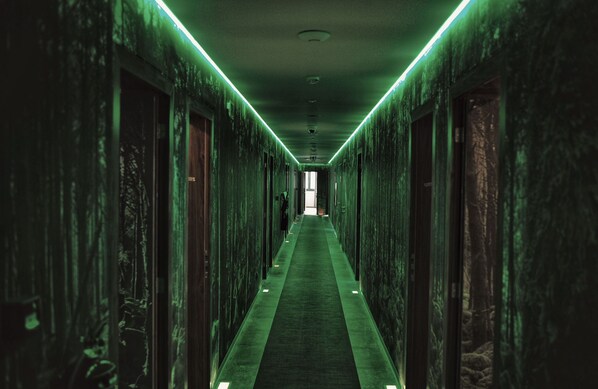 Un pasillo largo y estrecho con iluminación verde y suelo alfombrado, que conduce a una salida luminosa al final.