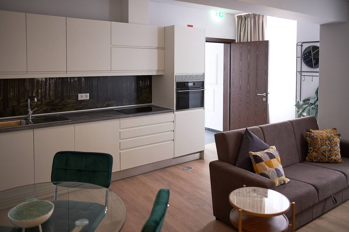 A modern kitchen with white cabinets, a black countertop, and a built-in oven. There is a dining area with a glass table and chairs, and a brown sofa with patterned cushions.