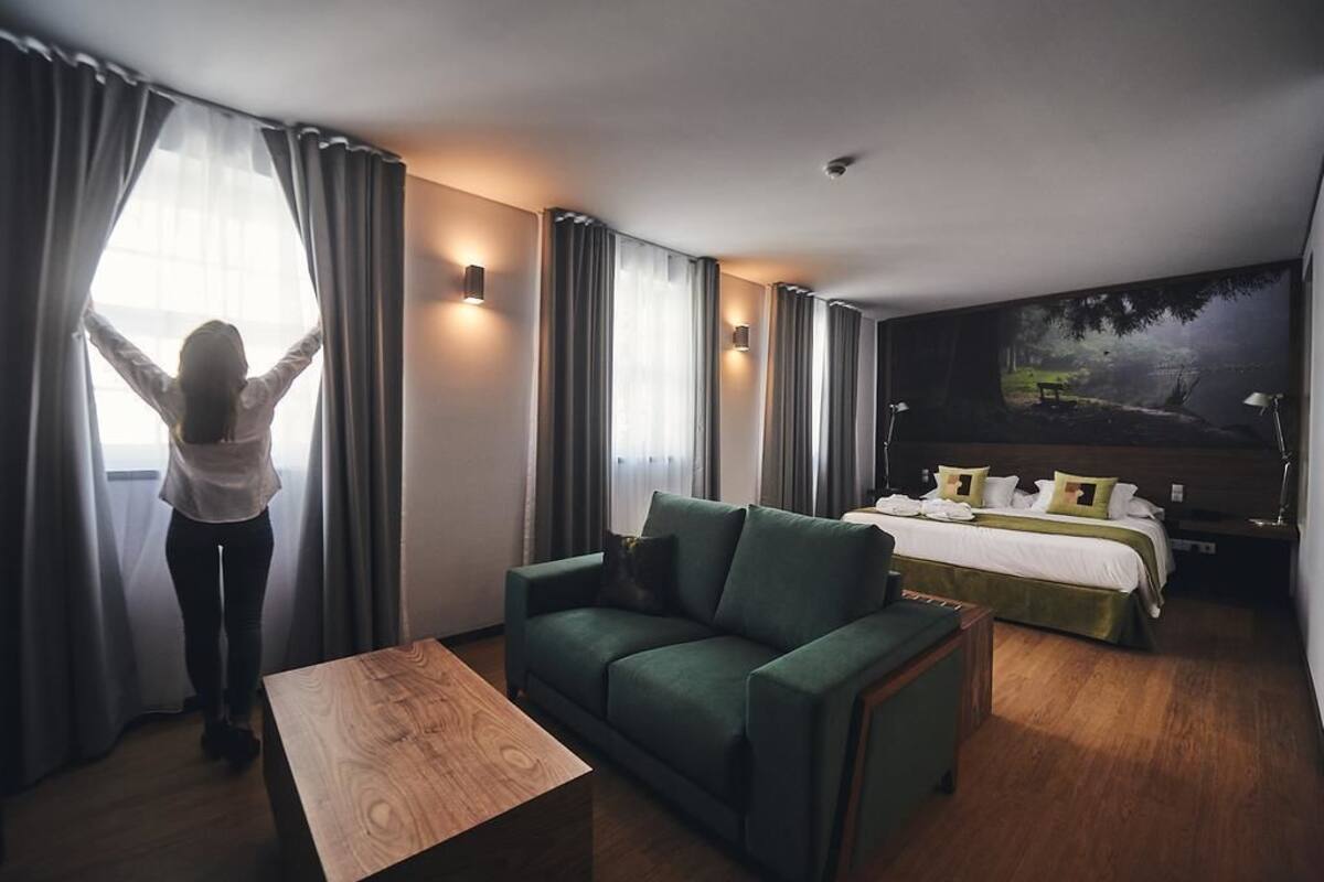 A hotel room with a green sofa, a wooden coffee table, a bed with white bedding, and a painting of a forest scene on the wall.