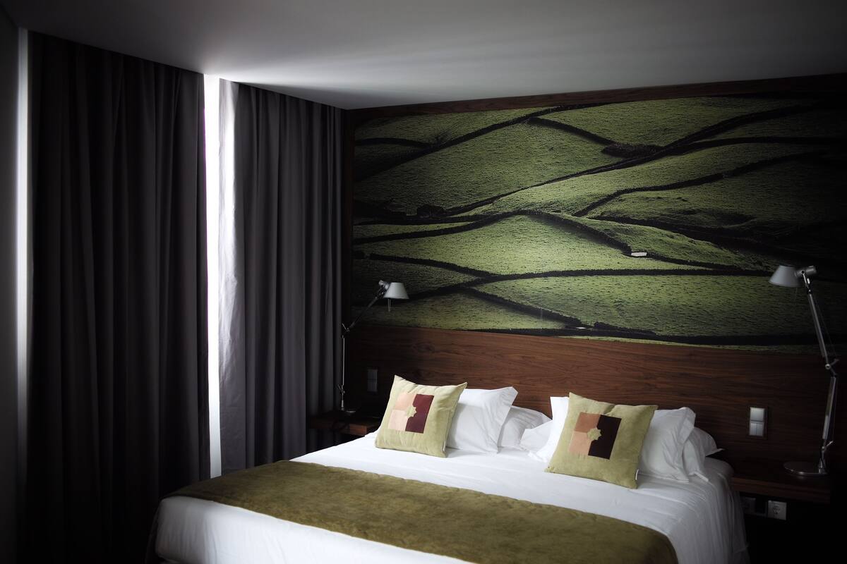 A hotel room with a large bed, a headboard with a tree branch design, a nightstand, a lamp, and a window with curtains.