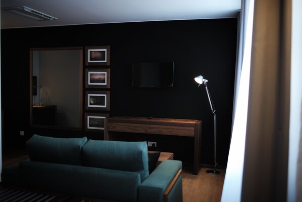 A dimly lit room with a teal sofa, a wooden desk, a standing lamp, and a flat-screen TV mounted on the wall.