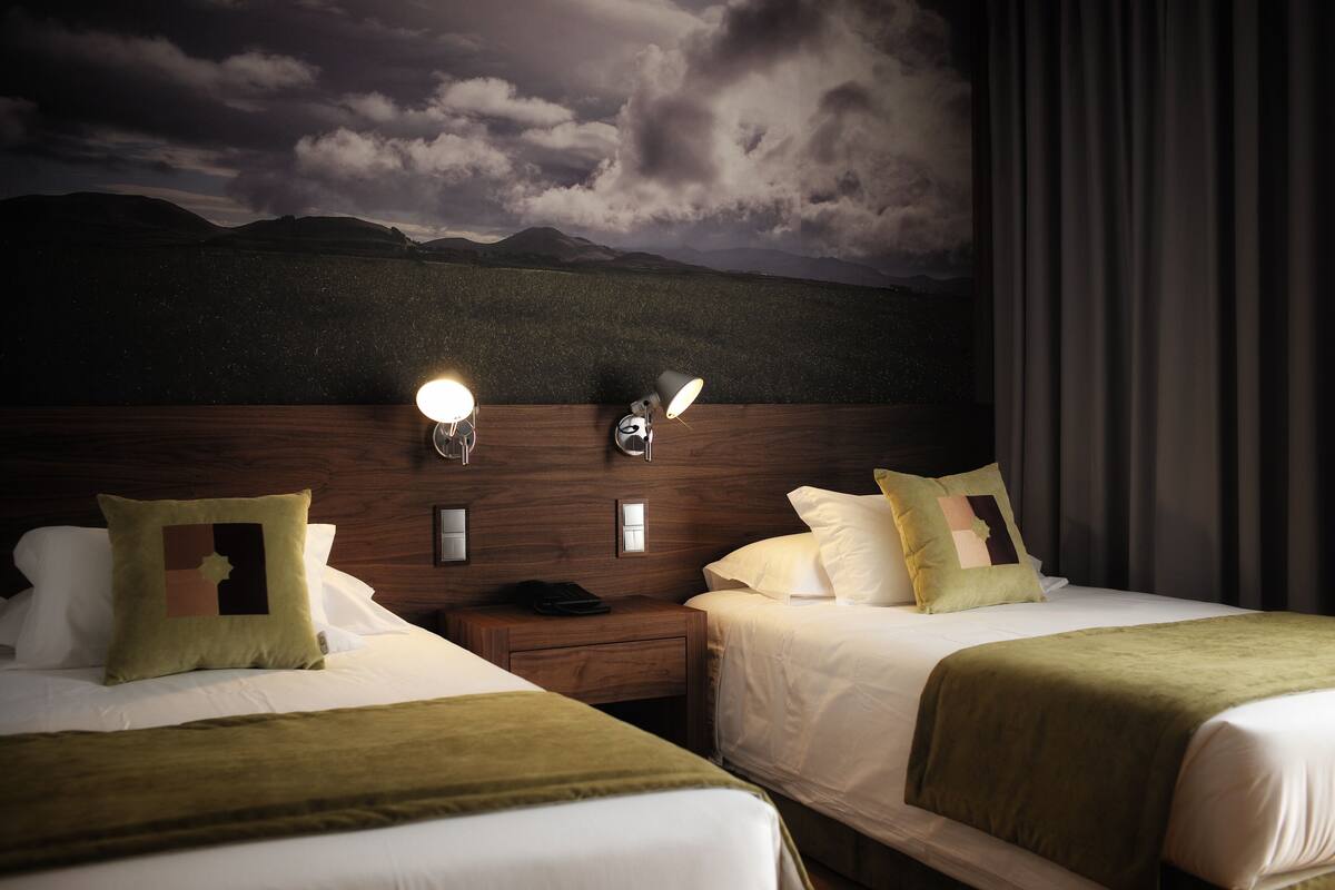 A hotel room with two beds, a nightstand, a lamp, and a painting of a mountain landscape.