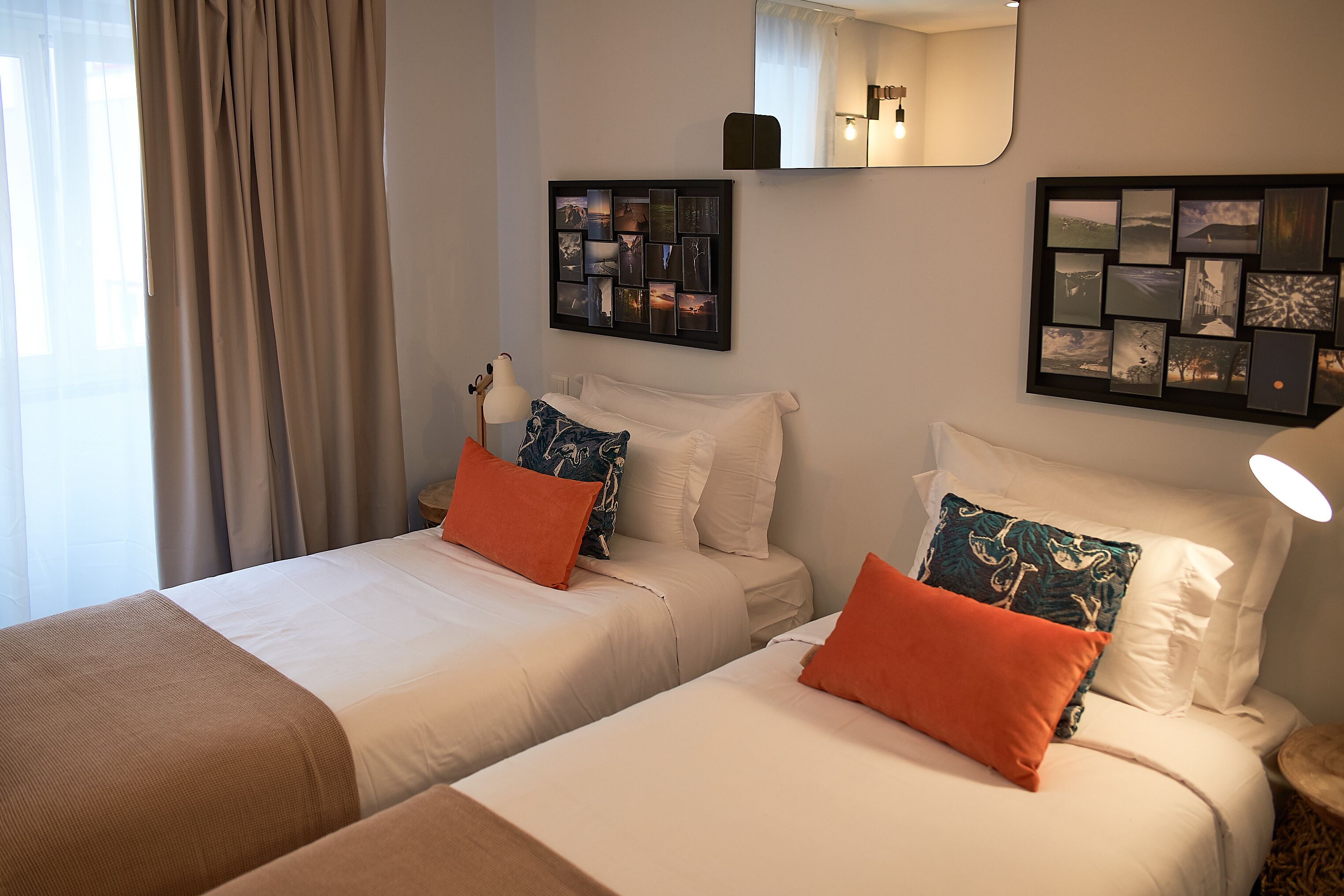 A hotel room with two beds, beige bedding, and orange pillows. There are wall-mounted picture frames and a mirror above the beds.